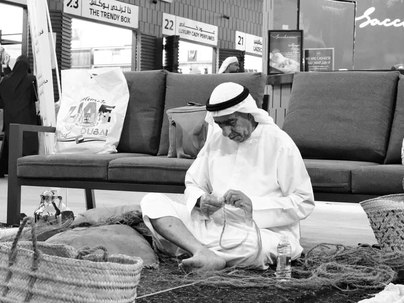 An elderly man in traditional attire sits on a carpet, weaving a basket in a market setting.