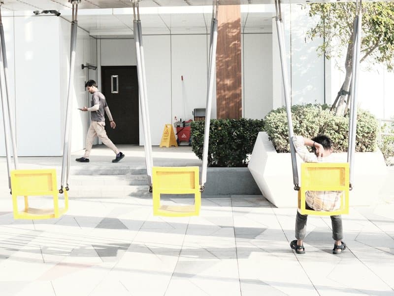 Two swings in a modern outdoor setting with a person walking by and another sitting on a swing.