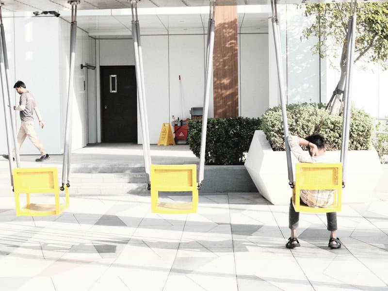 Two yellow swings in an urban setting with a person walking in the background and another sitting on a swing.