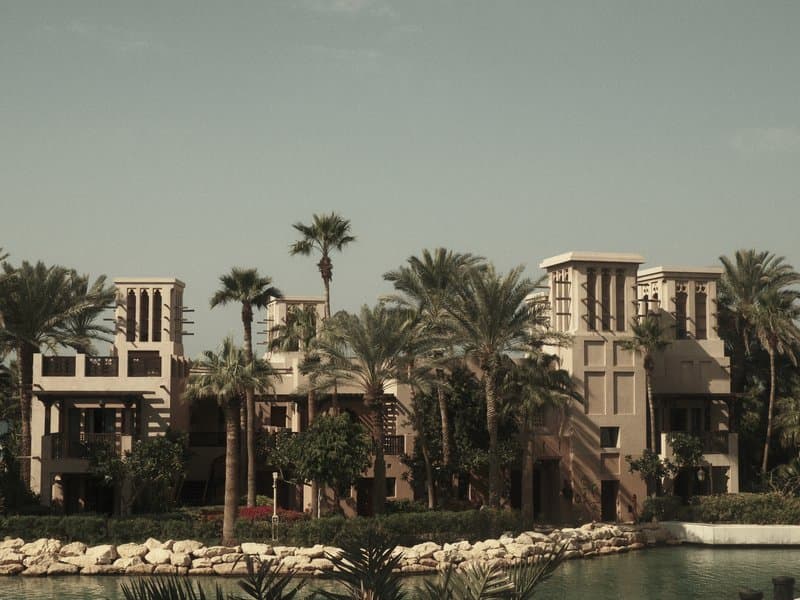 A modern building surrounded by palm trees and a calm water body under a cloudy sky.