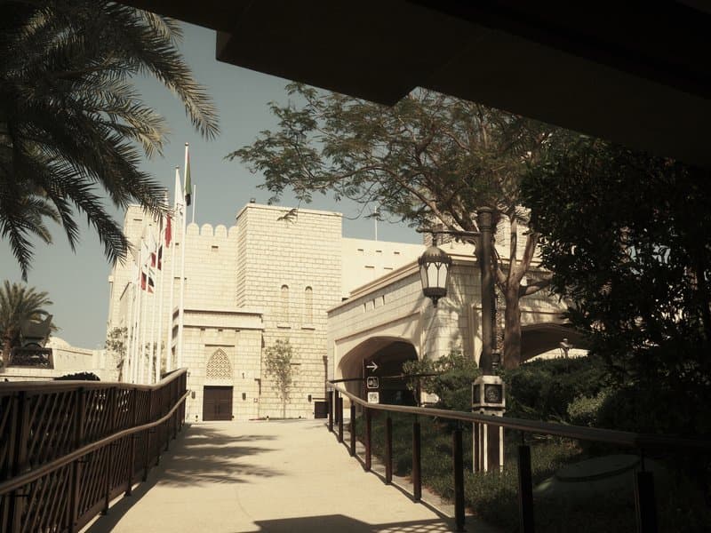 A historic building with palm trees and flags, viewed from a walkway under a shaded area.