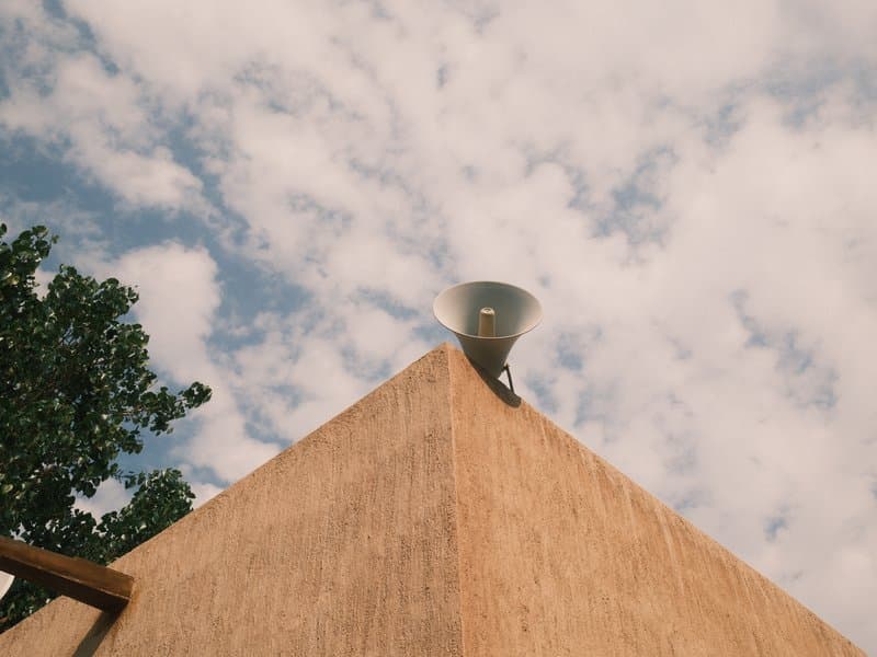 A close-up view of a building's corner with a speaker on top against a cloudy sky.