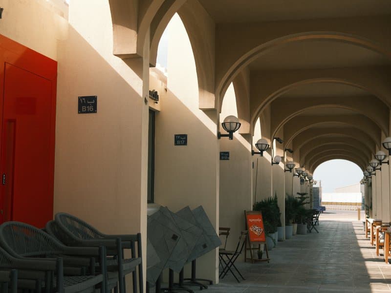 A corridor with arches and light fixtures, featuring empty chairs and a view of the sea in the distance.