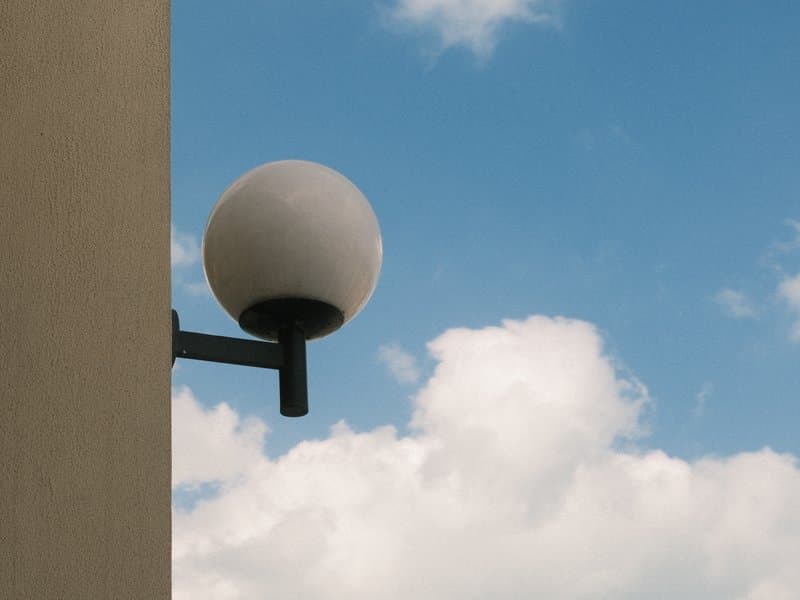 A round light fixture mounted on a wall against a blue sky with fluffy white clouds.