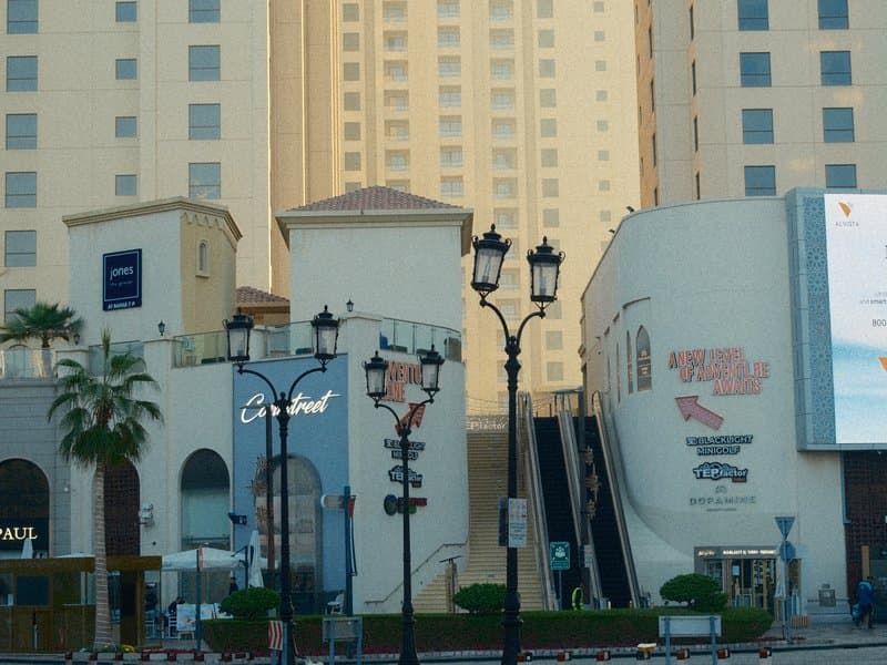 Urban scene featuring modern buildings, street lamps, and palm trees under soft evening light.