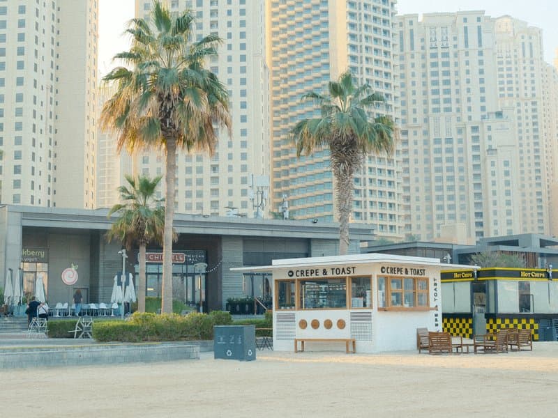 A beachside kiosk with palm trees and tall buildings in the background under soft light.