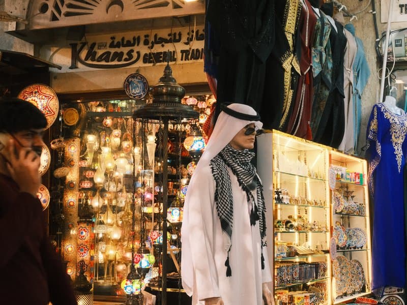 A man in traditional attire stands in a vibrant market filled with colorful lanterns and clothing.