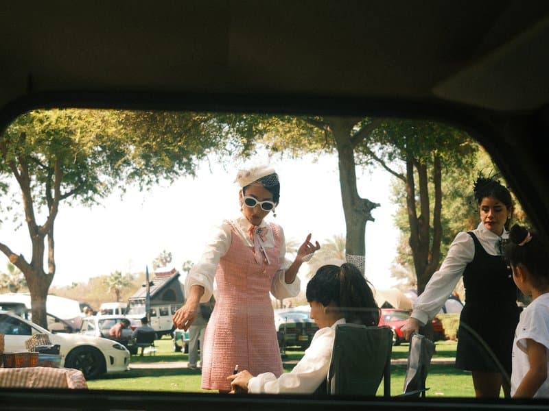 A woman in a pink dress gestures animatedly while another woman assists a child outdoors.