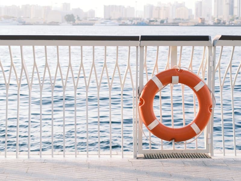 A bright orange life buoy hanging on a white railing with calm water and city skyline in the background.
