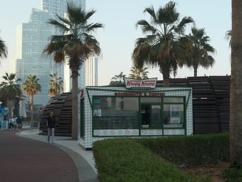 A small kiosk with a sign, surrounded by palm trees and modern buildings in the background.