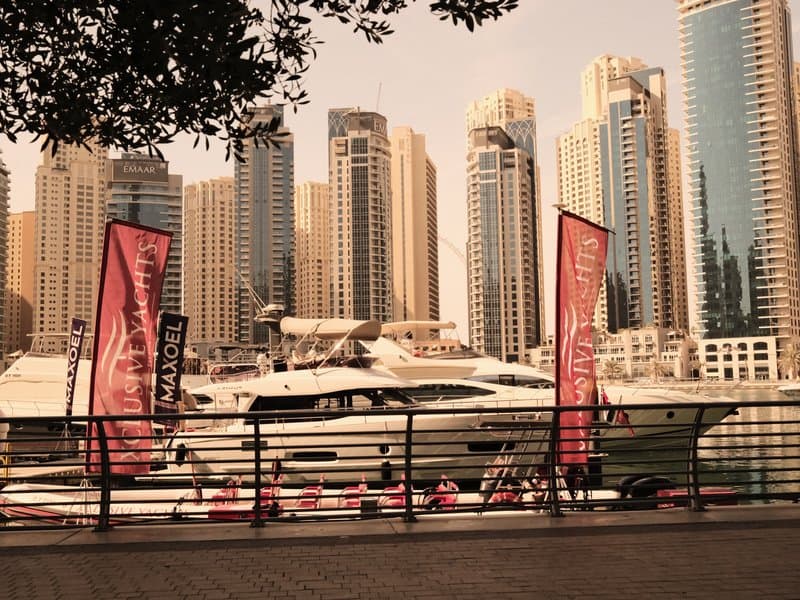 A luxury yacht docked by a waterfront with tall skyscrapers in the background and flags fluttering.