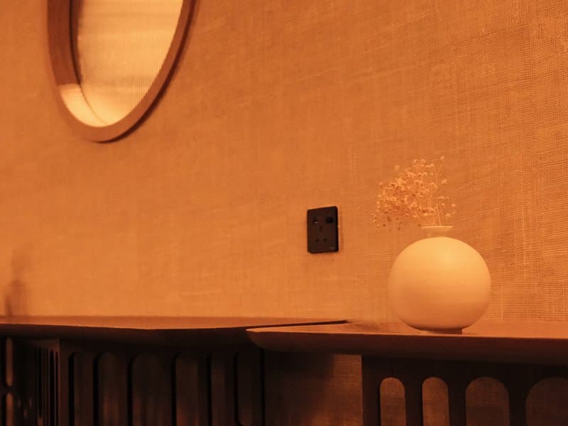 A minimalist interior scene featuring a round vase with dried flowers on a wooden shelf against a textured wall.