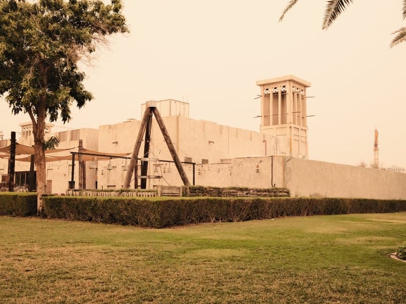 Historic building with a wind tower surrounded by green grass and palm trees under a hazy sky.