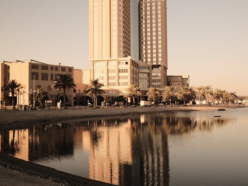 A tall building reflecting in calm water during golden hour, with palm trees lining the shore.