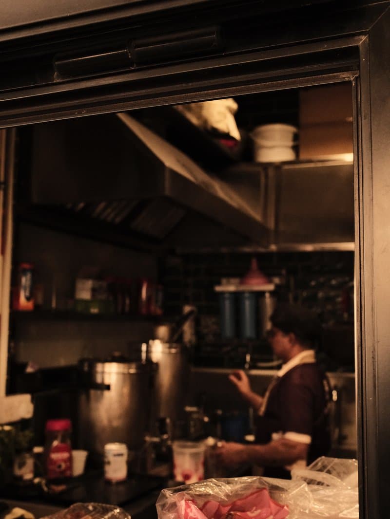 A dimly lit kitchen with a person preparing food, shelves filled with ingredients and utensils.