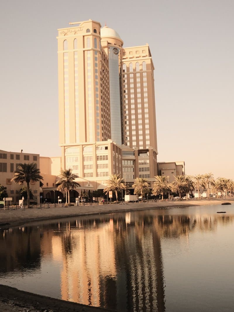 Tall buildings reflecting in calm water with palm trees in the foreground.