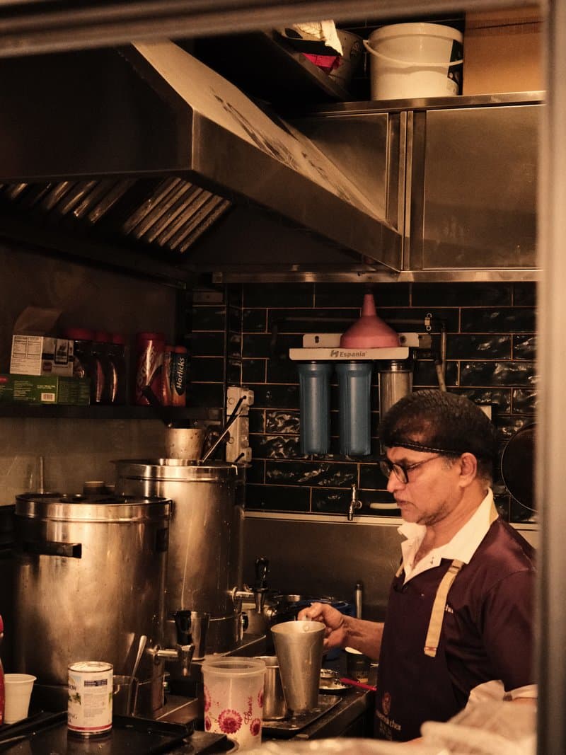 A barista preparing a drink in a dimly lit café kitchen with various equipment and containers around.
