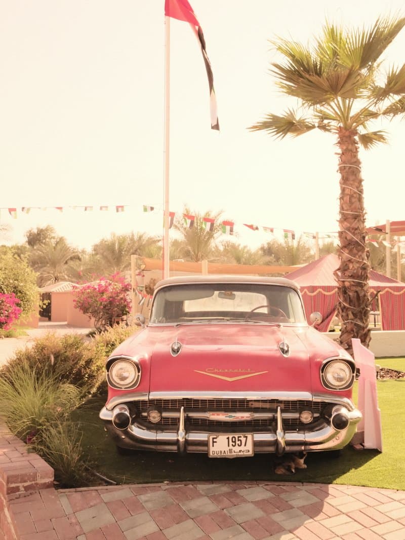 A vintage red car parked in a garden with palm trees and festive decorations.