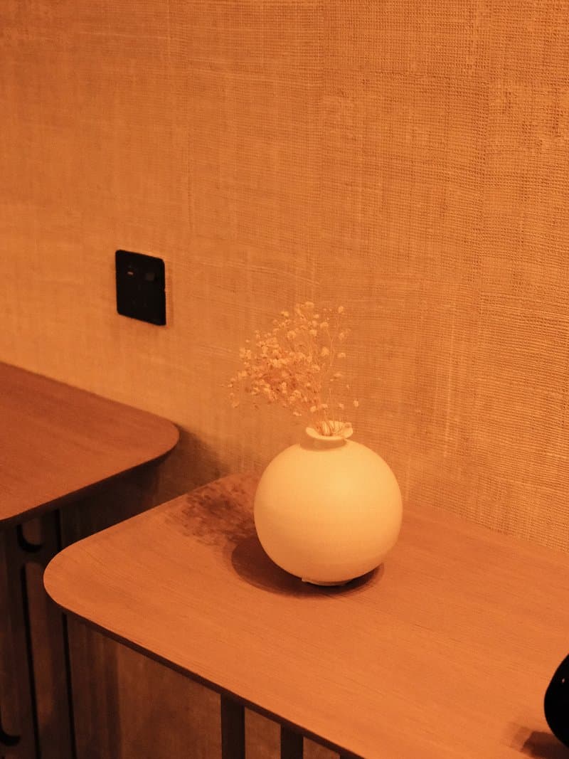 A minimalist white vase with dried flowers on a wooden table against a textured wall.