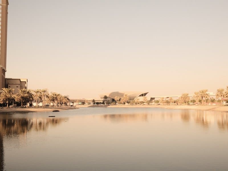 A serene river scene with reflections of buildings and trees under a soft pastel sky.
