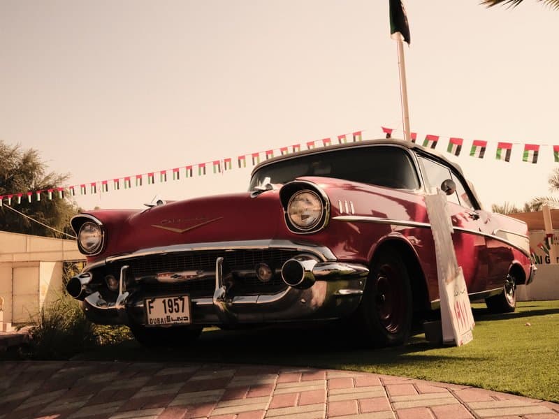 A vintage red car parked on a grassy area with decorative flags in the background under warm sunlight.