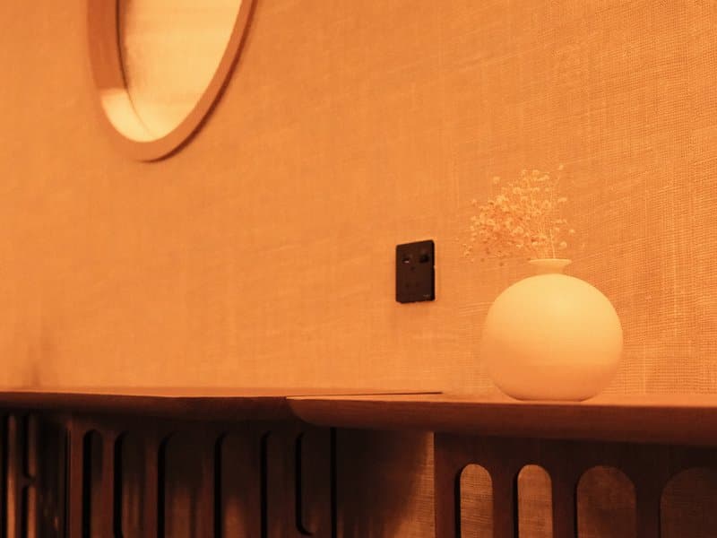 A round white lamp on a wooden shelf against a textured wall with a circular mirror.