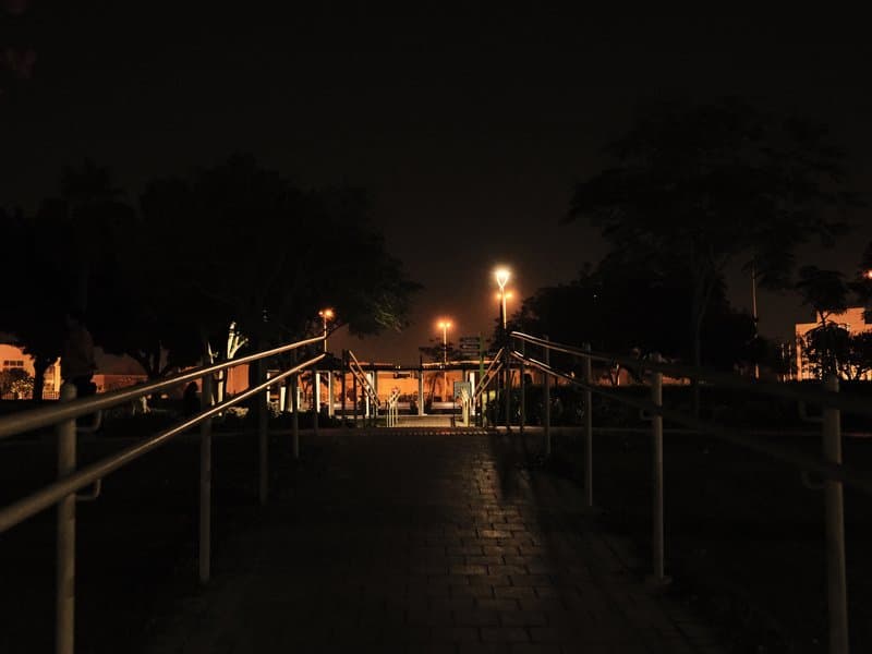 A dimly lit pathway with railings leading towards a distant light source in a park at night.