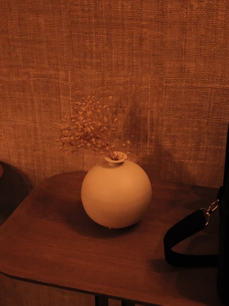 A simple white vase with dried flowers on a wooden table against a textured wall.
