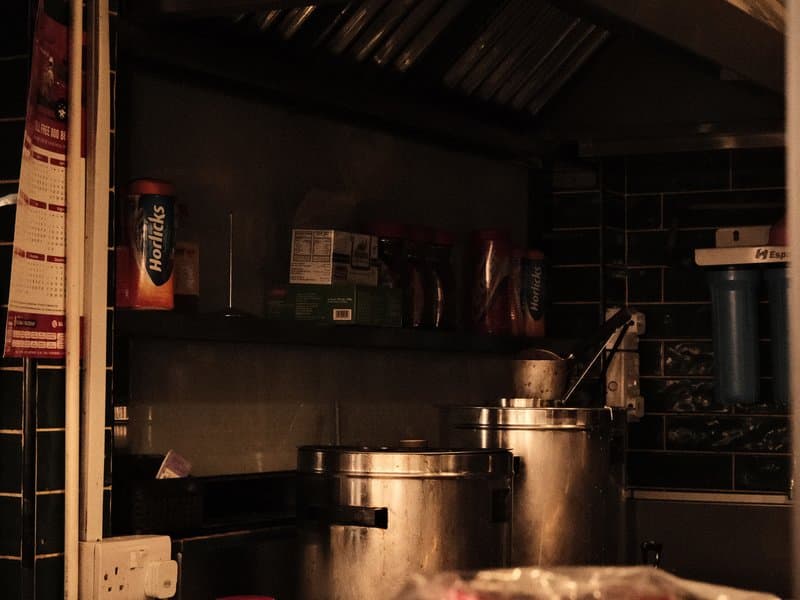 A dimly lit kitchen with metal pots on a shelf, various utensils, and a dark tiled wall.