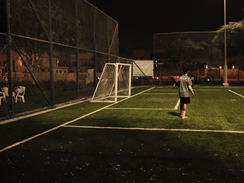 A person walking towards a soccer goal on a dimly lit field at night, surrounded by a fence.