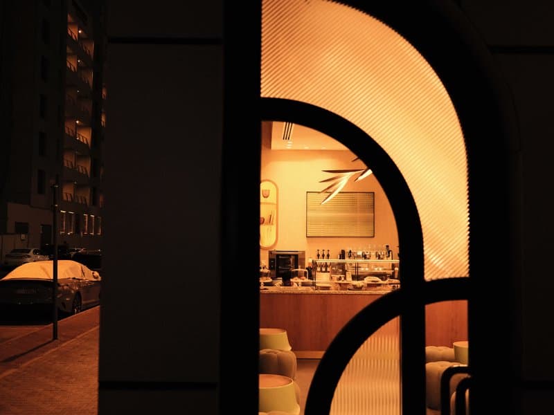 A warm-lit café interior viewed through an arched window at night, with soft seating and a bar area visible.