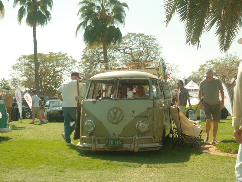 A vintage Volkswagen van parked on grass with people and palm trees in the background.