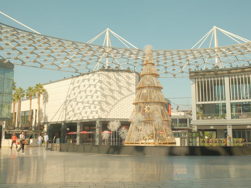 A large Christmas tree in an urban plaza with modern architecture and a patterned canopy overhead.