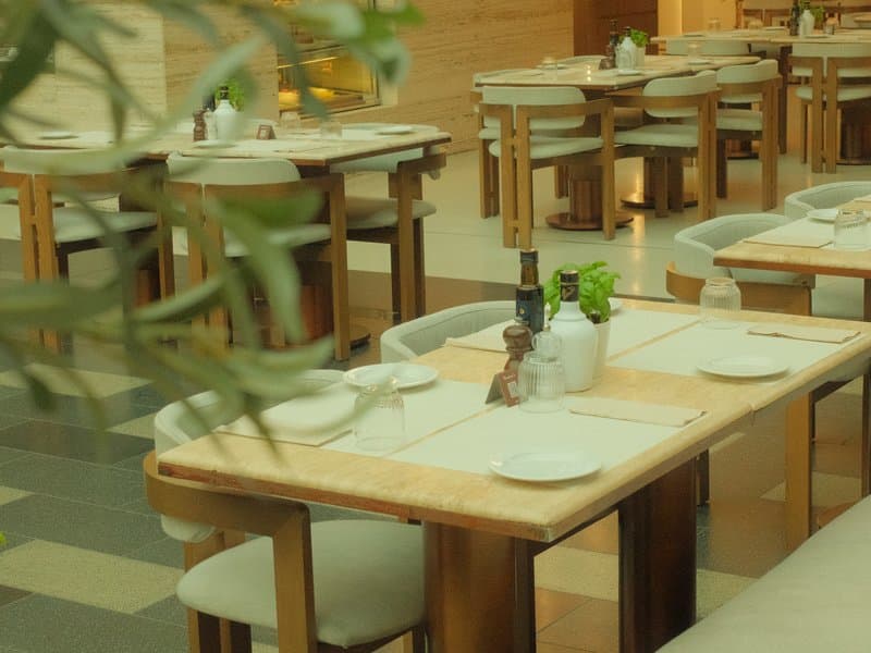 A modern restaurant interior with wooden tables and soft seating, plants in the background.
