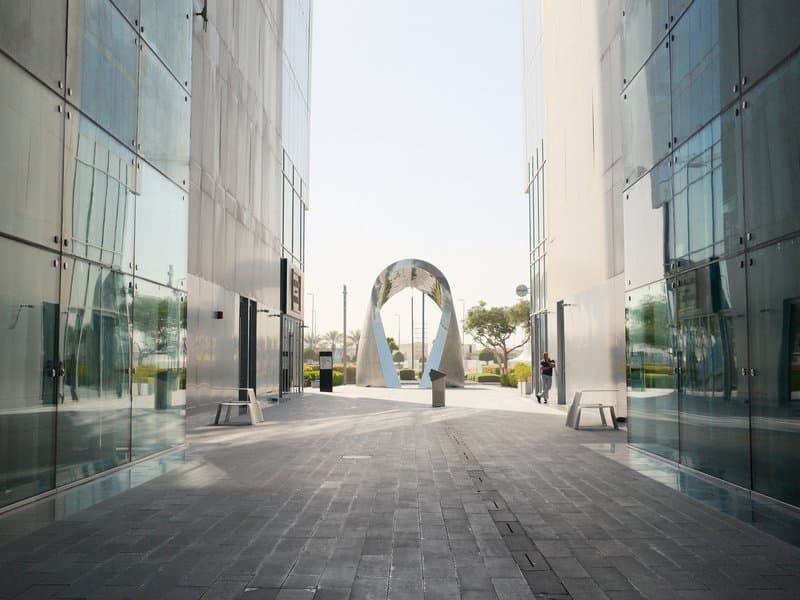 A modern urban pathway flanked by glass buildings leading to a sculptural arch.