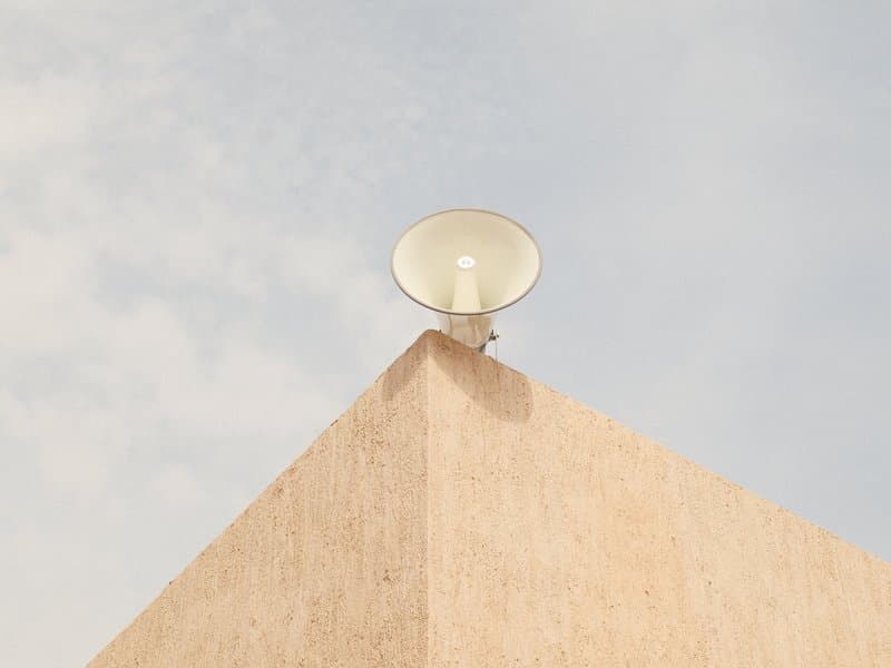 A large speaker mounted on a beige wall under a cloudy sky.