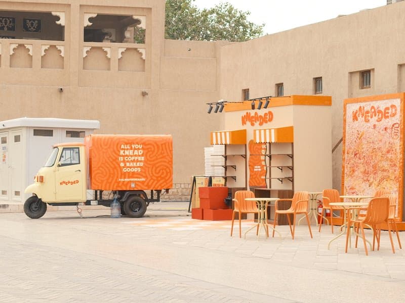 A colorful food truck and kiosk in a sandy urban setting with outdoor seating.