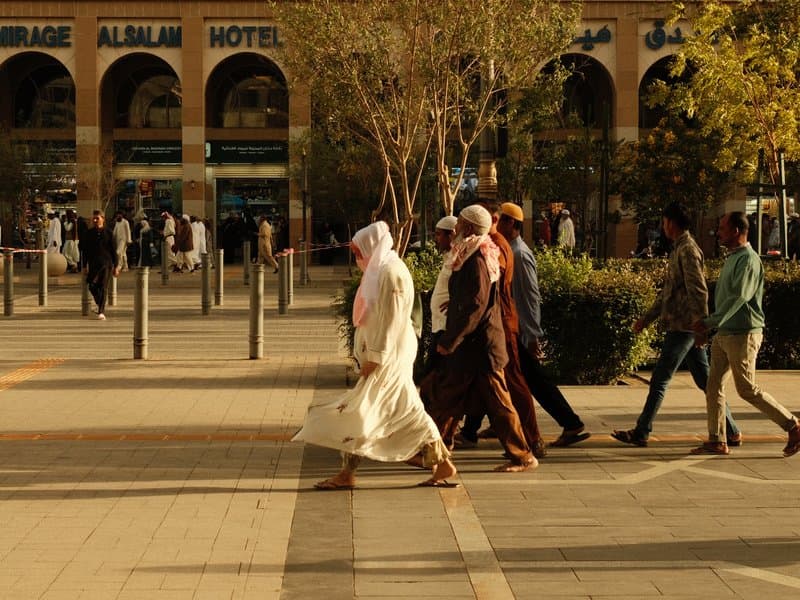 Group of people walking in a city plaza with trees and buildings in the background.