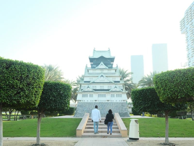 Two people walking towards a traditional building surrounded by manicured hedges and palm trees.