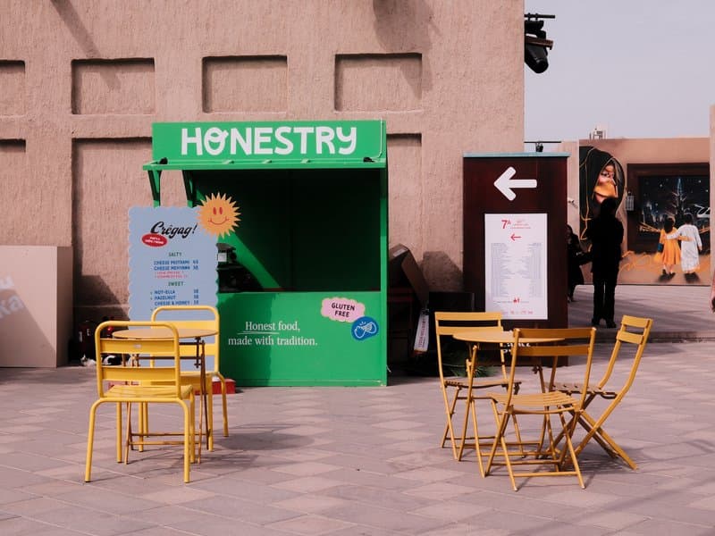A vibrant green kiosk labeled 'HONESTRY' with yellow chairs and tables in an urban setting.