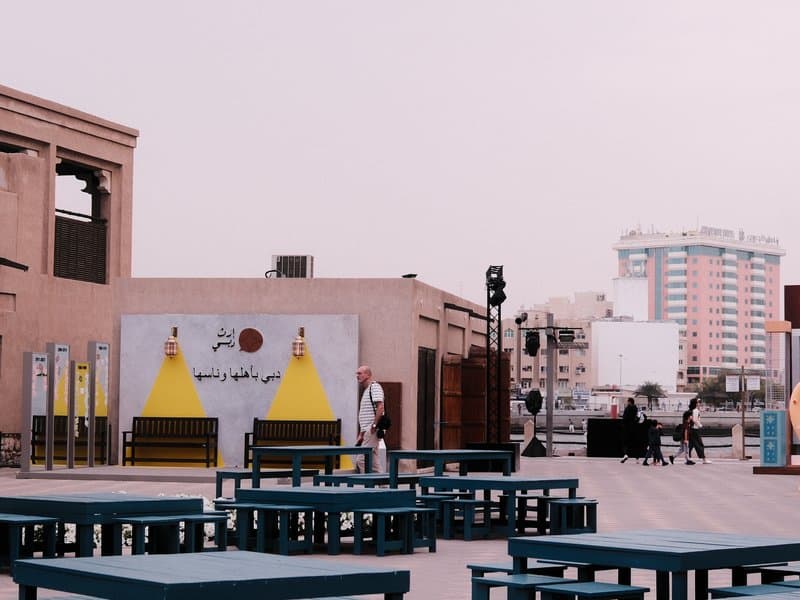 Outdoor urban scene with tables, a mural, and buildings in the background under a pinkish sky.
