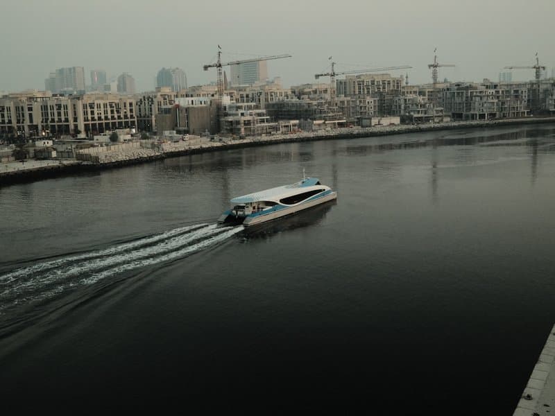 A boat gliding through a calm river with urban buildings and cranes in the background under a hazy sky.