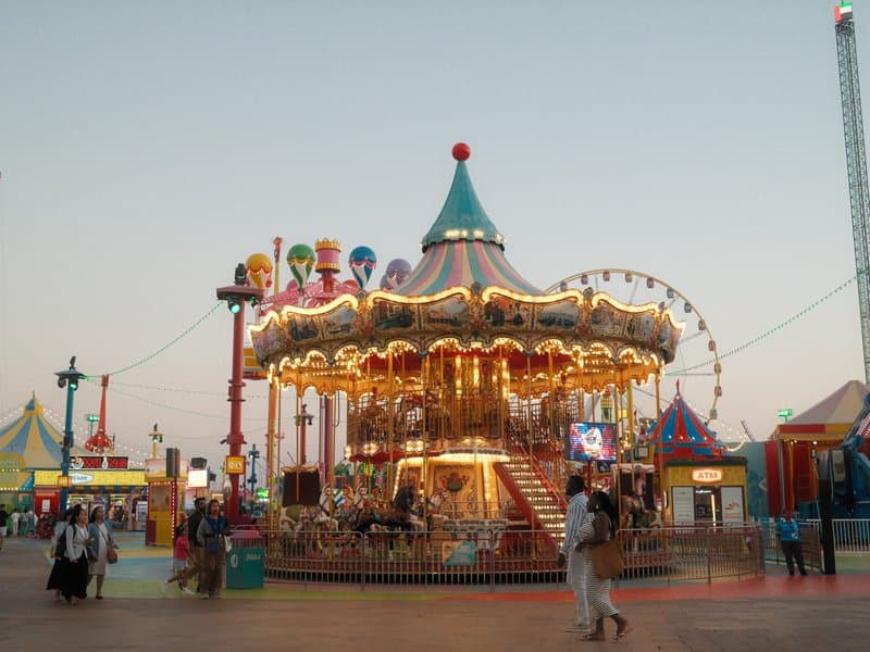 A brightly lit carousel at a fairground during twilight, surrounded by colorful tents and attractions.