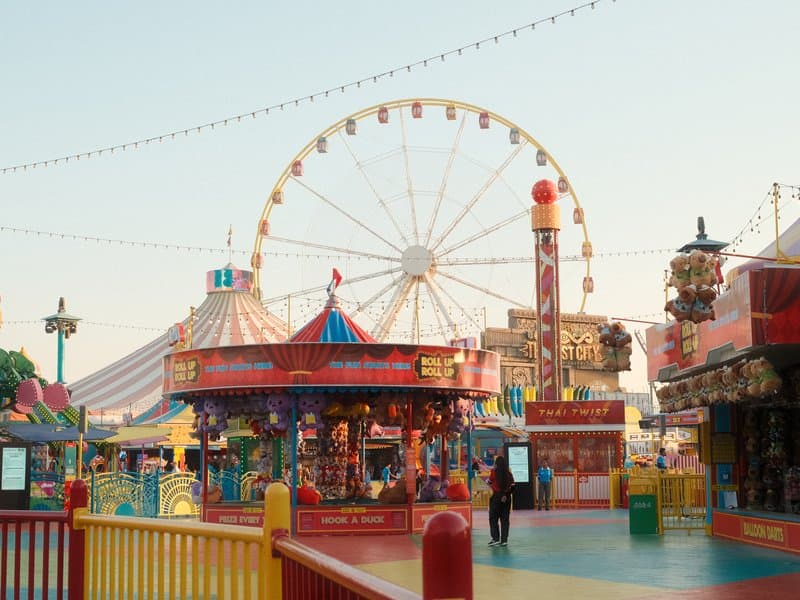 A vibrant amusement park scene featuring a ferris wheel, carousel, and colorful tents under a clear sky.