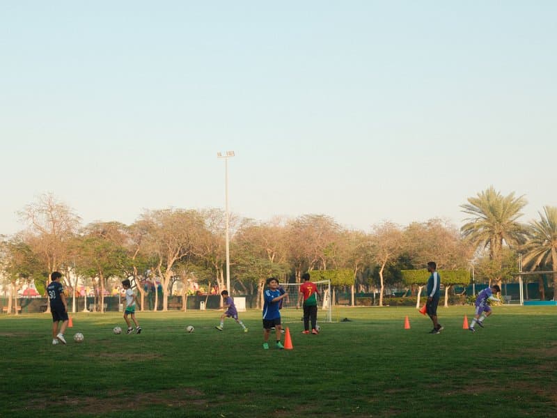 A group of people practicing soccer on a grassy field with trees in the background under a clear sky.