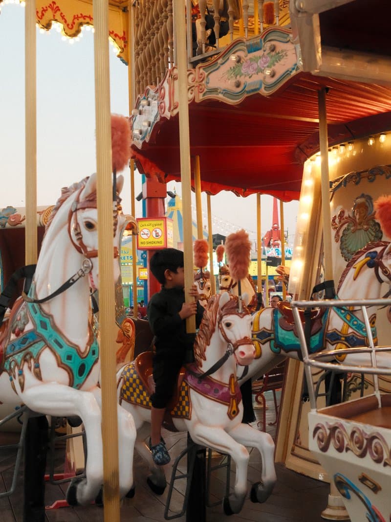 A child riding a carousel horse at a carnival, with colorful decorations and lights in the background.