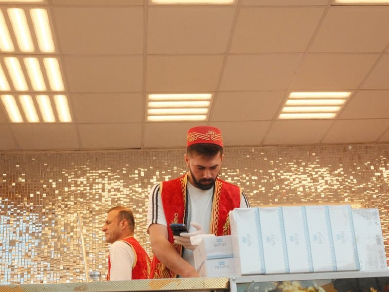 Two workers in red vests inside a bright shop, with a sparkling background and a counter filled with products.
