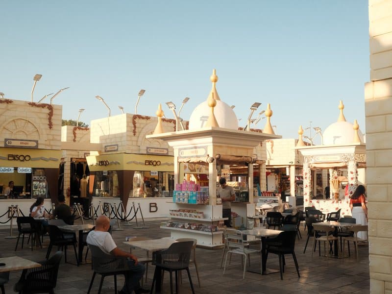 Outdoor market scene with colorful stalls and people seated at tables under a clear sky.