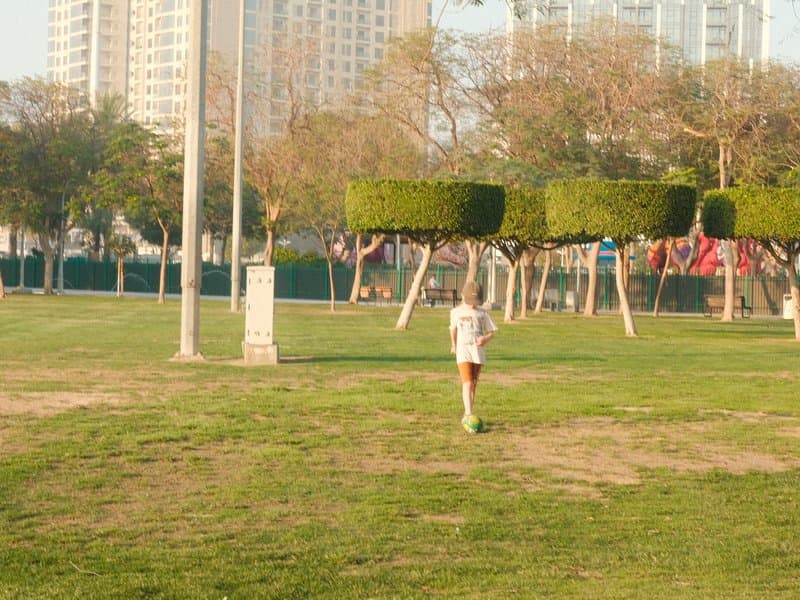 A child playing in a park with neatly trimmed bushes and tall buildings in the background.