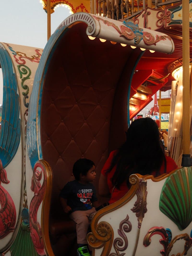 A child sitting on a colorful carousel seat with a parent nearby, vibrant carnival atmosphere.